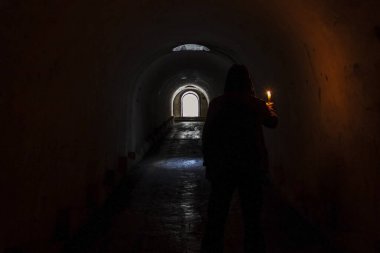 Kapüşonlu, elinde yanan bir mum tutan ve karanlık, kasvetli bir yeraltı tünelinin ışıklı çıkışında duran bulanık bir adam figürü..