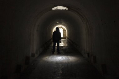 Kapüşonlu koyu renk elbiseli bir adam karanlık ve karanlık bir yeraltı tünelinden çıkan ışığa doğru gidiyor. Koridorun tavanındaki kapaktan güneş ışığıyla aydınlatılıyor..
