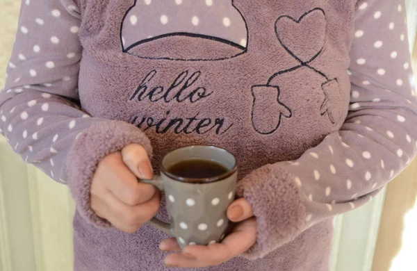 Winter is cold. A woman in warm pajamas holds a cup of hot tea in her hands.