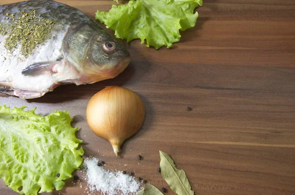 On the table lies a fresh, gutted river fish, crucian carp, onions, lettuce, salt, spices.