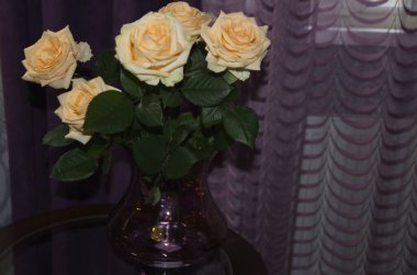 Mor tekstil arka planında, güzel bir vazoda yuvarlak cam bir masanın üzerinde yeşil yapraklı güzel kokulu turuncu güller var..