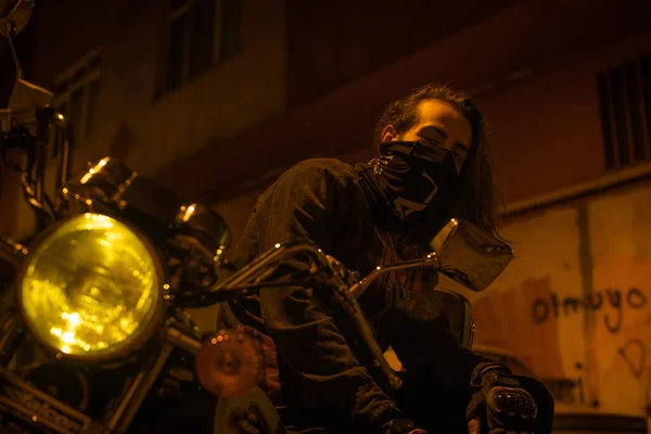 Young man with long hair mask on motorcycle on night street