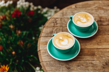 Yeşil seramik bardakların güzel üst manzarası taze kahveyle dolu latte sanatıyla, ahşap masa üzerinde servis edilen çiçeklerle süslenmiş..
