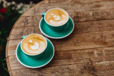 Yuvarlak ahşap masa, iki mavi seramik fincan lezzetli, kalp şeklinde süt köpüğünden yapılmış kahve. Aroma içeceği. Sabah uyanmak için taze demlenmiş..