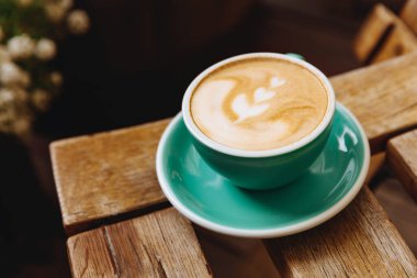Yeşil seramik fincanın üzerinde güzel latte resimli, ahşap masa üzerinde, arka planda bulanık fincan tabağı olan kahve bardağını kapat. Profesyonel barista müşteri için lezzetli bir içecek yaptı.