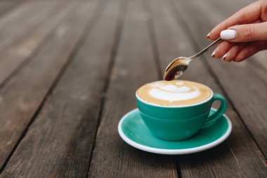 Beyaz manikürlü kadın eli sıcak taze aroma kapuçinosunun üzerinde kaşıkla açık mavi bardakta latte sanatı. Genç güzel kız sabah içkisinin tadını çıkaracak.