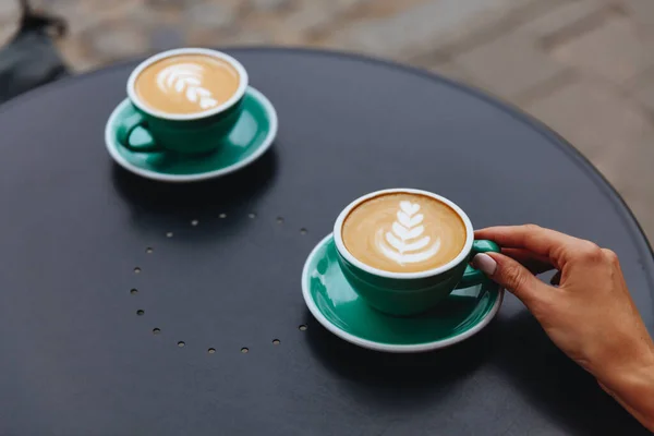 Woman hand holding one out of two light blue cup of hot aroma capuccino with lush foam milk served on black table. Young lady waiting to her friend to enjoy morning hot drinks together outdoors.