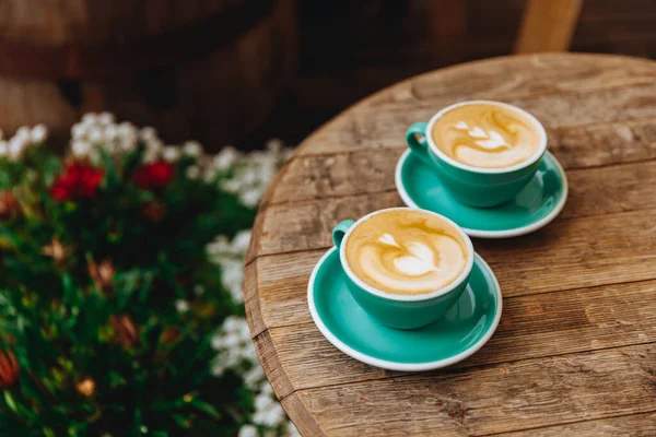 Açık mavi kahve fincanları, çiçek çalılarının bulanık arkaplanlı ahşap masa üzerinde kalp şeklinde latte sanatıyla birlikte. Retro, vintage efekti ve kopyalama alanı