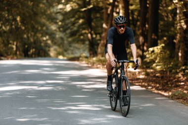Spor kıyafetleri ve siyah kasklı bisiklet giyen profesyonel bisikletçi yaz ormanları arasında asfalt yolda bisiklet sürüyor. Koruyucu gözlüklü sakallı sporcu dışarıda antrenman yapıyor..