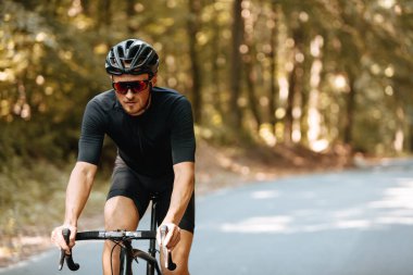 Spor kıyafetleri, koruyucu kaskı ve güneş gözlüğü takan sakallı bir adam asfalt yolda siyah bisiklet sürüyor ve geçmişi bulanık. Sıkı eğitim ve öz disiplin kavramı