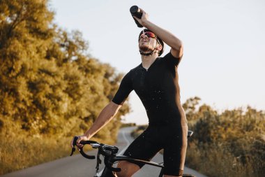 Spor elbiseli, koruyucu kasklı ve aynalı gözlüklü olgun bir adam bisikletle kaldırımlı yolda dururken tatlı su içiyor. Tazeleme ve etkinlik kavramı