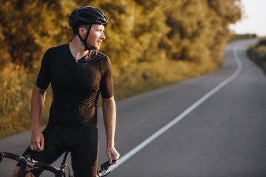 Spor elbiseli, siyah kasklı, sakallı bir adam motosikletinin üzerinde oturuyor ve kaldırımlı yol ve yeşil ağaçların bulanık arka planına bakıyor. Faaliyet ve motivasyon kavramı.