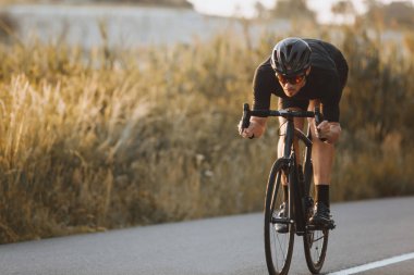 Asfalt yolda siyah spor kıyafeti, kask ve gözlük takan aktif profesyonel bisikletçi. Güçlü adam becerilerini geliştiriyor ve bisiklet yarışmasına hazırlanıyor.