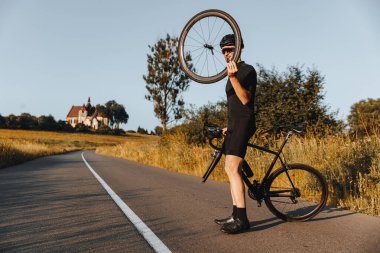 Aktif giyimli, koruyucu kasklı ve aynalı gözlüklü, kaldırımlı yolda kırık bisikletli olgun bir adam. Sakallı bisikletçi elinde tekerlekle arabasını tamir etmeye çalışıyor..