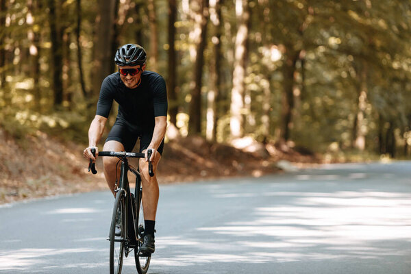 Happy bearded person in protective helmet, mirrored glasses and activewear biking on paved road in green forest during summer days. Concept of regular training and healthy lifestyle.