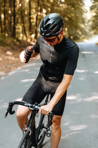 Professional cyclist in protective helmet and eyeglasses stopped in green forest to drink fresh water. Mature man in activewear taking rest during riding.