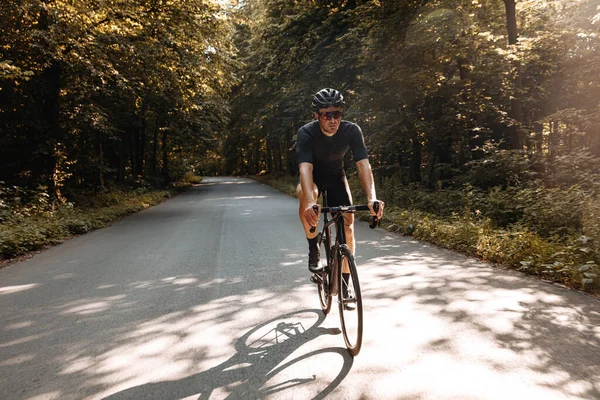 Active bearded athlete in sport clothing riding bike on paved road during sunny days. Strong sportsman in protective helmet and glasses training regularly outdoors.