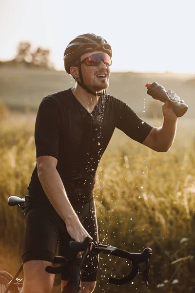 Strong bearded man in active wear, protective helmet and sport glasses splashing fresh water from bottle outdoors. Tired cyclist feeling thirsty after hard workout.