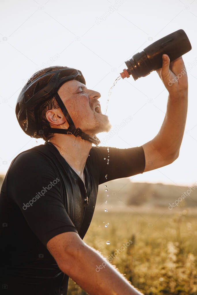 Side view of exhausted bearded sportsman in black helmet pouring water from sport bottle on his face. Mature man refreshing after long distance riding.
