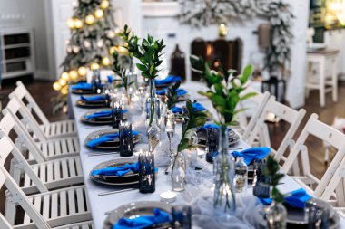 Lüks tabaklarla, parlak çatal bıçaklarla, bardaklarla ve mavi peçetelerle servis edilen uzun masa. Noel arifesi için şenlik dekorasyonu. Şöminesi ve Noel ağacı olan güzel bir oturma odası..