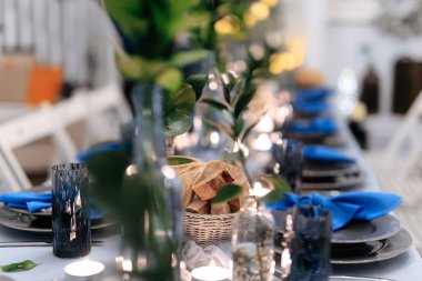 Lüks tabaklı, parlak çatal bıçaklı ve mavi peçeteli şenlik masasının bulanık arka planı. Mumlar, kumaşlar ve yeşil yapraklardan yapılmış güzel bir dekorasyon..