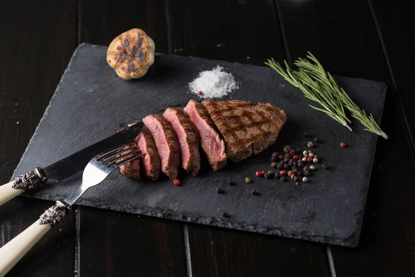 Filete de ribeye de primera calidad en rodajas en placa de piedra negra. Grado medio de cocción del filete. Con romero y pimientos. Tenedor y cuchillo . —  Fotos de Stock