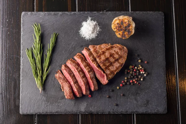 Bife de ribeye em fatias na placa de pedra preta. Grau médio de doneness de bife. Com alecrim e pimentos. Garfo e faca . — Fotografia de Stock