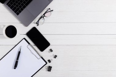Düz yatıyordu, ahşap ofis masasının üstünde. Not defteri, dizüstü bilgisayar, akıllı telefon, not panosu, ataç, ataç, gözlük, kahve fincanı ofis malzemesi ve beyaz ahşap masa arkaplanı.