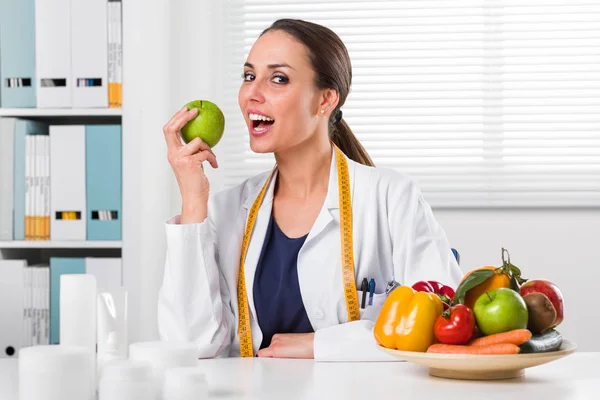 Улыбающаяся женщина-диетолог ест зеленое яблоко в своем офисе — стоковое фото