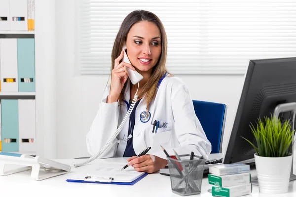 Sonriente doctora sentada en el escritorio con teléfono al oído —  Fotos de Stock