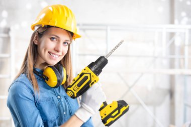 Genç bayan inşaatçı, yeni beyaz bir duvarı delmeye çalışıyor. Pille çalışan bir matkapla kameraya bakıyor ve gülümsüyor. 
