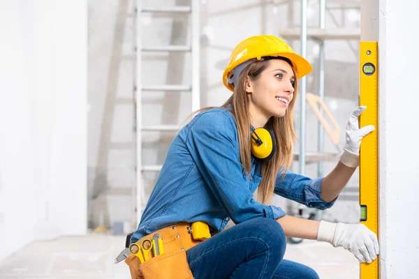 Attractive young female construction worker in yellow hardhat checking level of the wall with the spirit level tool. Close-up portrait