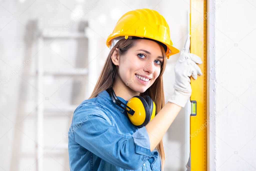 Attractive young female construction worker in yellow hardhat checking level of the wall with the spirit level and turning to smile at camera 