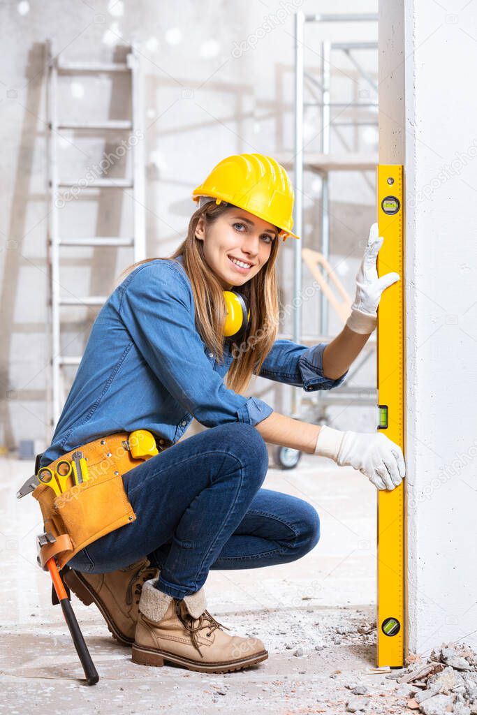 Attractive young female construction worker in yellow hardhat checking level of the wall with the spirit level and turning to smile at camera 