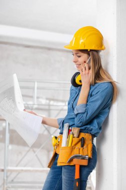 İnşaat halindeki bir odada akıllı telefonuyla sohbet ederken elinde bir plan tutan genç kadın mühendis ya da mimar. 
