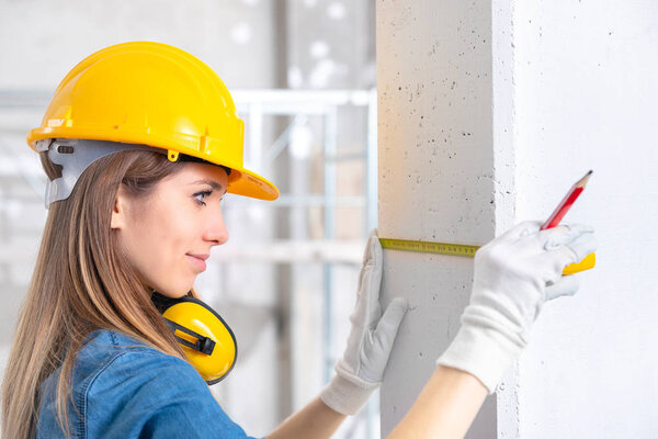 Young female construction worker in yellow hardhat and gloves measuring the wall with measuring tape. Indoor portrait 