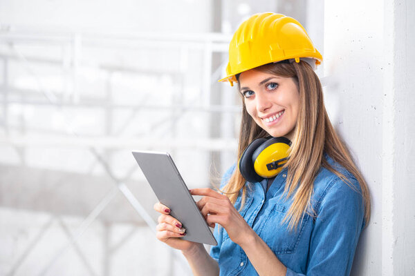 Smiling young female construction worker wearing a hardhat and ear muffs using a tablet computer on site on a high key background