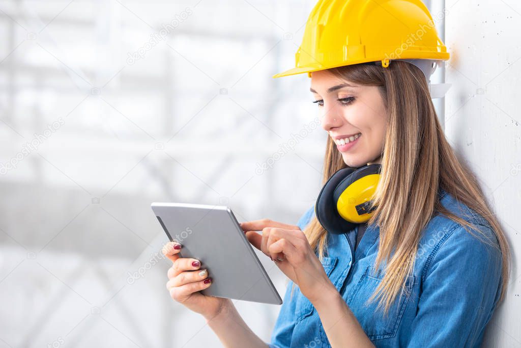 Smiling young female construction worker wearing a hardhat and ear muffs using a tablet computer on site on a high key background