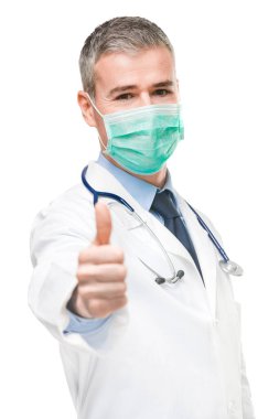 Cerrahi yüz maskesi, laboratuvar önlüğü ve steteskop takan bir doktor, Corona virüsü ya da Covid-19 konsepti üzerine odaklanmış bir şekilde başparmak kaldırıyor.
