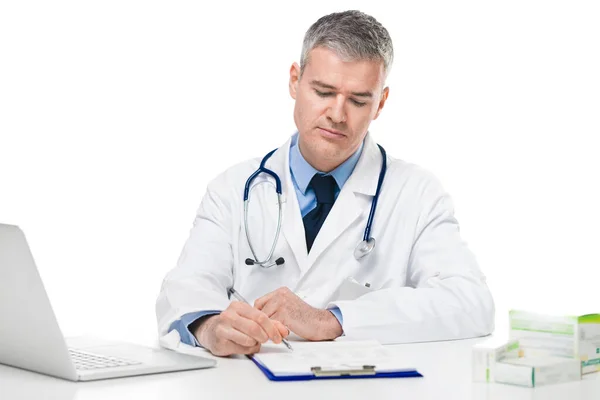 白い背景の上に彼の仕事に集中彼のオフィスで患者のメモや処方箋を書いて座っている白い研究室のコートの医者 — ストック写真