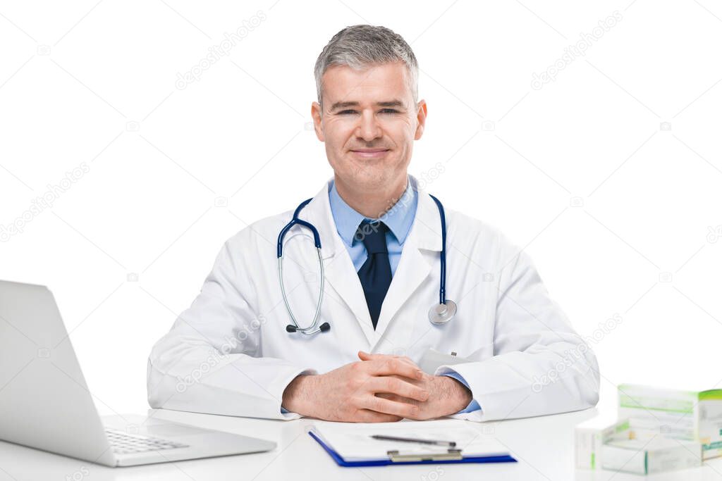 Successful confident doctor seated at a desk in his office behind a laptop computer looking at the camera with a quiet smile against a white background