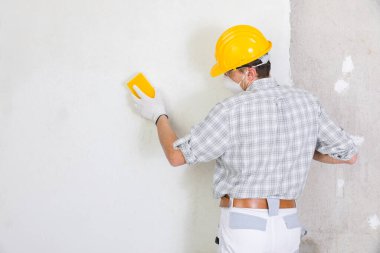 İnşaatçı zımparalıyor ve düzleştiriyor. Yeni sıvanmış beyaz bir iç duvarı evin içinde. Toz maskesi ve şapka takarak boyamaya hazırlanıyor.