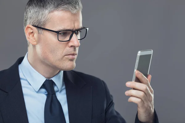 Stylish businessman watching to a mobile phone screen with a serious thoughtful expression, head and shoulders isolated on grey
