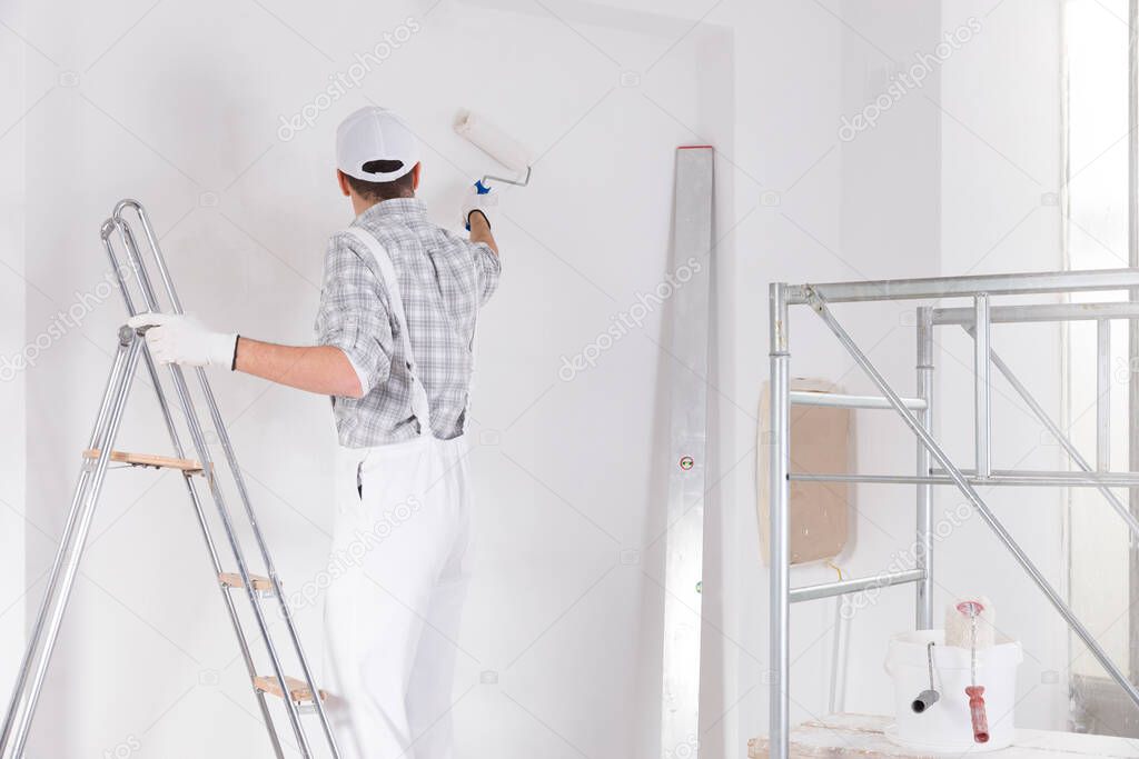 Painter standing on a ladder painting a white wall in a room under construction leaning away using a roller viewed from the rear with interior scaffolding frame alongside