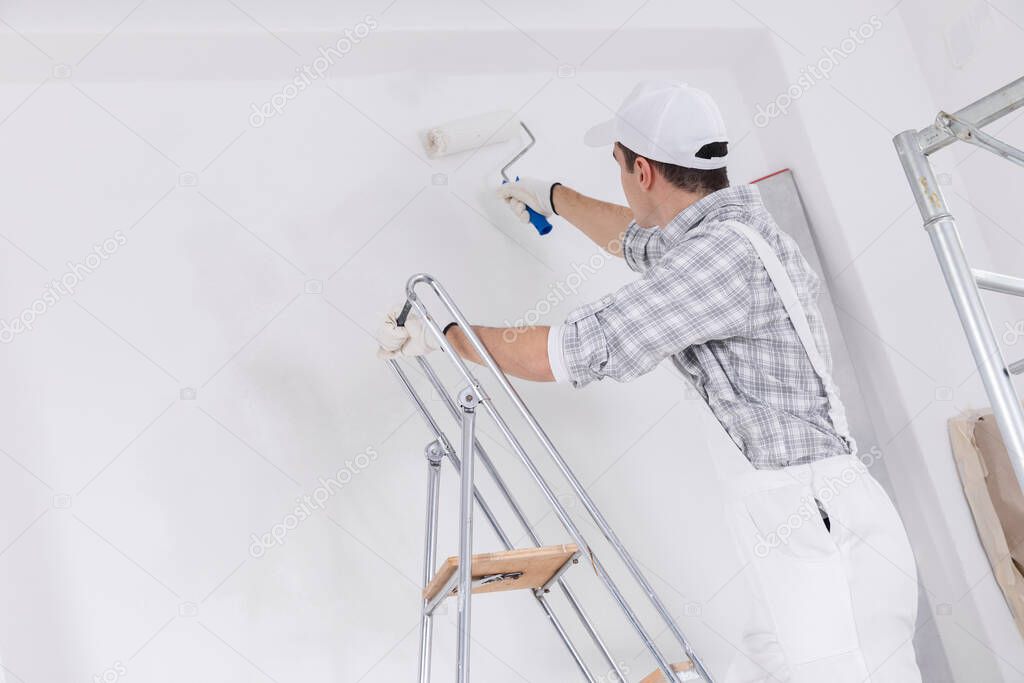 Painter or builder painting a white wall standing on a ladder using a roller in closeup looking away as he works