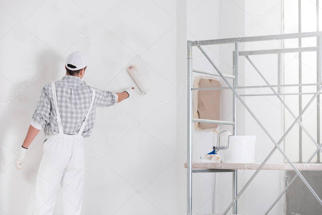 Painter or builder painting a wall with white paint and a roller viewed from behind with a ladder alongside in a DIY, renovation or construction concept