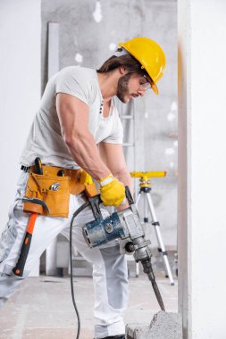 İnşaat halindeki bir odanın içindeki beton blokta kriko çekici kullanan bir inşaat işçisi kameraya gülümsüyor.