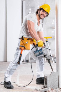 İnşaat halindeki bir odanın içindeki beton blokta kriko çekici kullanan bir inşaat işçisi kameraya gülümsüyor.