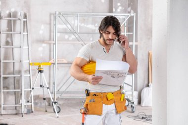 Tamamlanmamış bir odada, cep telefonunda fotokopi aletiyle sohbet ederken elinde bir plan tutan yakışıklı mimar ya da mimar.