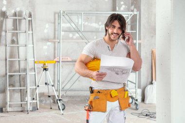 Gülümseyen mimar ya da mimar tamamlanmamış bir odada cep telefonunda fotokopi aletiyle sohbet ederken elinde bir plan tutuyor.